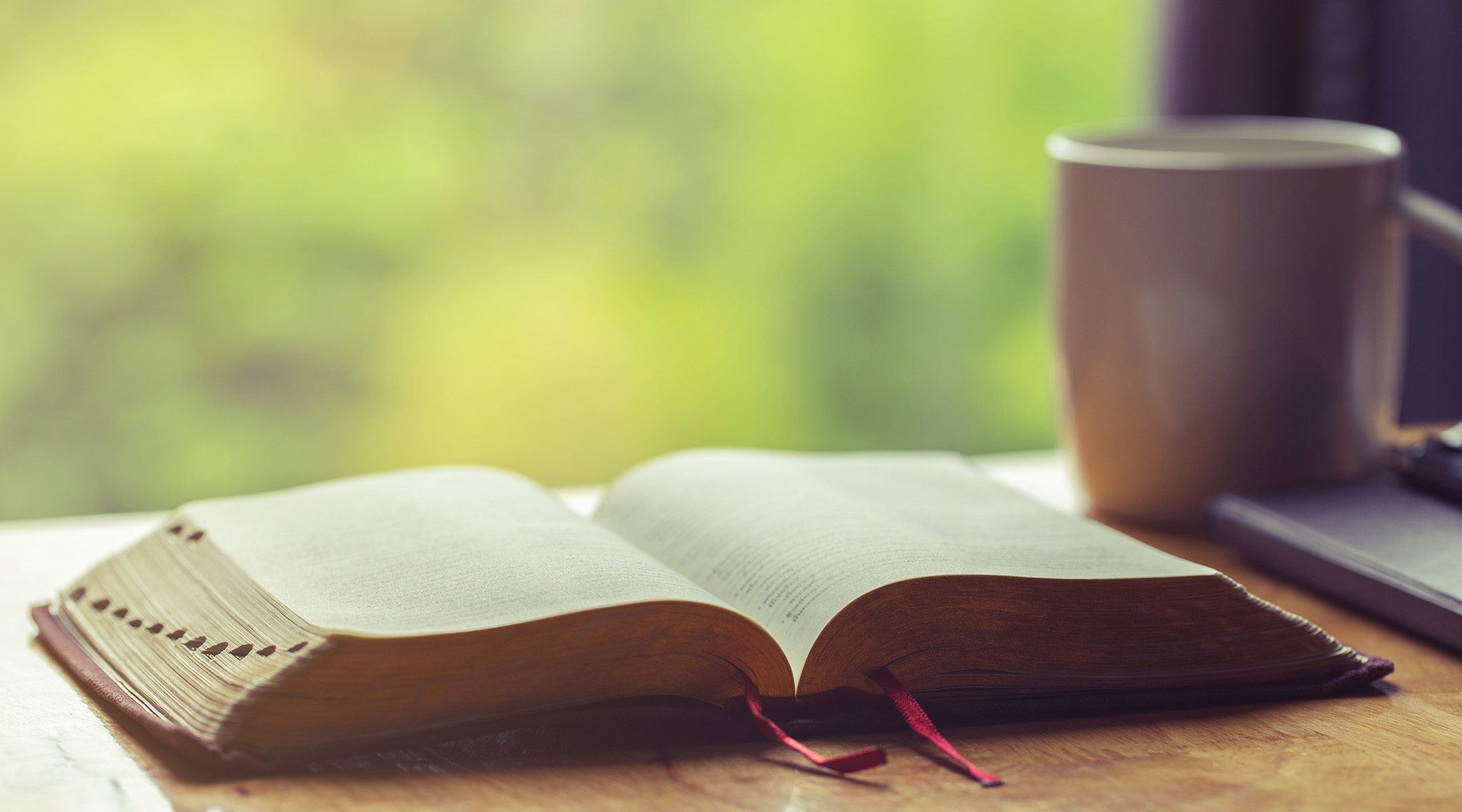 An open Bible on a table with a bookmark halfway through, warm morning light